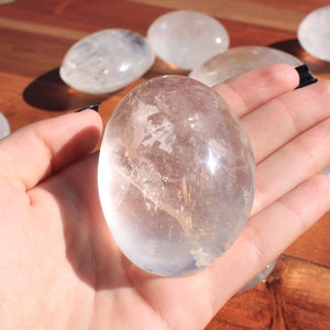 May include: A clear quartz crystal egg, polished smooth, with a translucent appearance. The egg is held in a hand, showcasing its size and shape.
