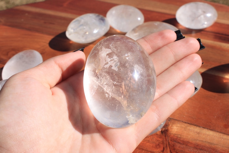 May include: A clear quartz crystal egg, polished smooth, with a translucent appearance. The egg is held in a hand, showcasing its size and shape.