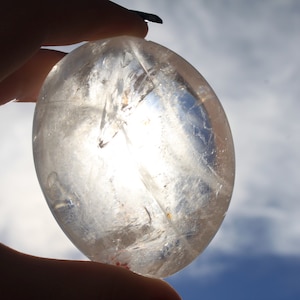 May include: A clear quartz crystal sphere, with a smooth, polished surface, held in a hand against a cloudy blue sky.
