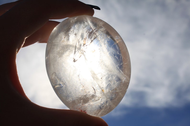 May include: A clear quartz crystal sphere, with a smooth, polished surface, held in a hand against a cloudy blue sky.