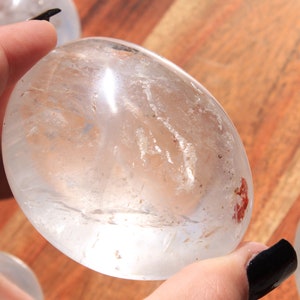 May include: A clear quartz crystal egg with a smooth, polished surface. The egg is translucent and has a slight milky hue. It is approximately 2 inches in diameter.