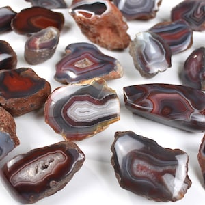 May include: A collection of polished agate stones in shades of red, gray, white, and brown. The stones vary in size and shape, showcasing natural patterns and banding. Some stones have a rough, textured exterior.