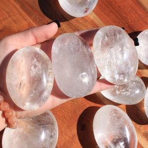 May include: A collection of eight clear quartz crystal palm stones. The stones are smooth and polished, with a slightly oval shape. They are arranged on a wooden surface.