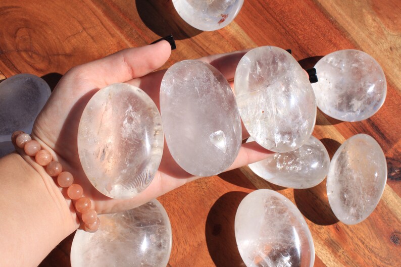 May include: A collection of eight clear quartz crystal palm stones. The stones are smooth and polished, with a slightly oval shape. They are arranged on a wooden surface.