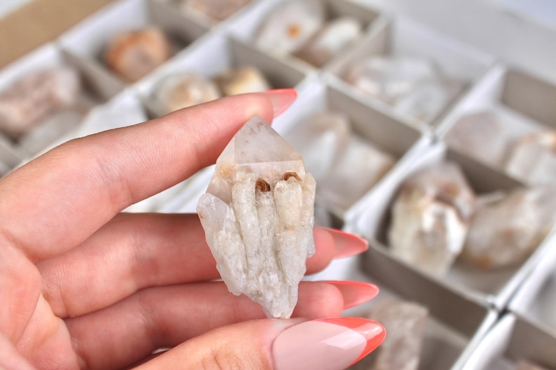 May include: A clear quartz crystal with a pointed tip and a rough, textured surface. The crystal is held in a hand with manicured nails, with other crystals in the background. The crystal is approximately 2 inches in length.