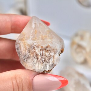 May include: A clear quartz crystal point with a natural, rough texture. The crystal has a pointed tip and a base with a mix of white, brown, and tan colors. The crystal is held in a hand.