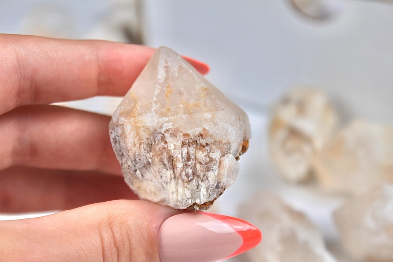 May include: A clear quartz crystal point with a natural, rough texture. The crystal has a pointed tip and a base with a mix of white, brown, and tan colors. The crystal is held in a hand.