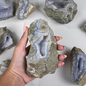 May include: A collection of blue lace agate geodes with a light blue interior and a rough, gray exterior. One geode is held in a hand. The geodes vary in size and shape, with visible crystal formations.