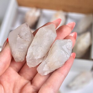 May include: Three clear quartz crystal points are held in a hand with pink nail polish. The crystals are rough-cut and have a translucent appearance. Other crystals are visible in a box in the background.