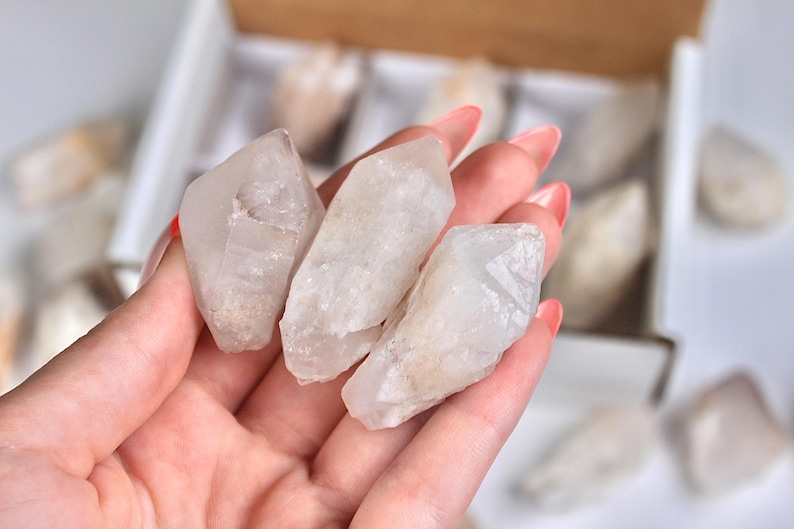 May include: Three clear quartz crystal points are held in a hand with pink nail polish. The crystals are rough-cut and have a translucent appearance. Other crystals are visible in a box in the background.