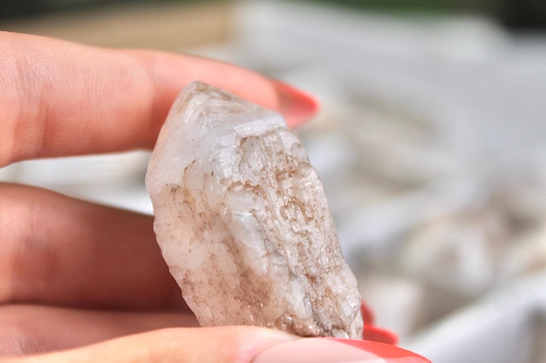 May include: A rough, translucent quartz crystal with a natural, textured surface. The crystal is held between fingers, showcasing its irregular shape and subtle color variations, ranging from clear to light brown.