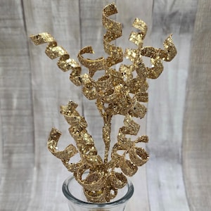 以下が含まれることがあります： 透明なガラスの花瓶に飾られた、スパイラルリボンの付いた金色の装飾的な茎。茎は茶色のベースで、フラワーアレンジメントやホリデーデコレーションに最適です。花瓶は明るい木目調の表面に置かれています。