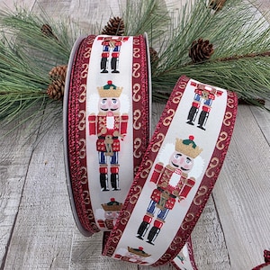 May include: A roll of decorative ribbon featuring a repeating pattern of a toy soldier nutcracker on a white background. The ribbon has a burgundy border with a gold swirl design. Pine needles and pine cones are in the background.