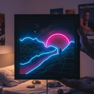 May include: A framed artwork with a neon landscape design. The image shows a person on a mountain, with a pink sun setting behind the mountains. The mountains and sun are outlined in neon blue, creating a striking visual.