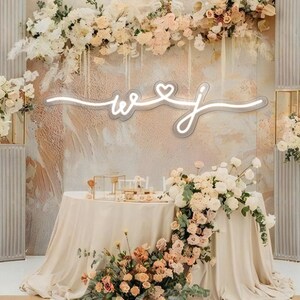 May include: A wedding ceremony backdrop with white and cream floral arrangements, two tall white columns, and a neon sign that reads "W & J" with a heart in the middle.