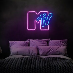 May include: A neon sign with the MTV logo in pink and blue. The sign is mounted on a black wall above a bed with purple pillows and a gray knitted blanket.
