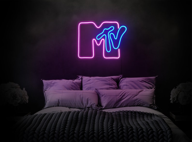 May include: A neon sign with the MTV logo in pink and blue. The sign is mounted on a black wall above a bed with purple pillows and a gray knitted blanket.