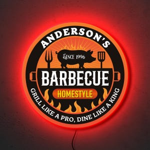 Pode incluir: Uma placa redonda vermelha iluminada com o texto "Anderson's Barbecue" em branco, "Homestyle" em um oval vermelho e "Grill like a pro, dine like a king" em preto. A placa também apresenta um porco, chamas e ferramentas de churrasco.