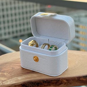 Puede incluir: Una pequeña caja de joyería blanca con un cierre dorado, que contiene tres anillos. Un anillo es dorado con un diseño de estrella, otro tiene pequeñas piedras y el tercero tiene piedras verdes. La caja está sobre una tabla de madera.