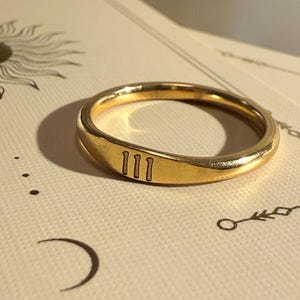 Peut inclure: Une bague dorée avec le chiffre "111" gravé sur le devant. La bague est posée sur une surface crème avec des illustrations sur le thème céleste, dont un soleil, un croissant de lune et d'autres symboles.