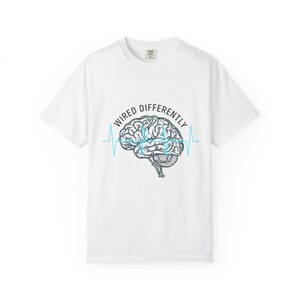 以下が含まれることがあります： 白いTシャツ。脳のグラフィックと「WIRED DIFFERENTLY」という青い文字がプリントされています。脳のイラストは青いアクセントと青い心拍ラインで詳細に描かれています。