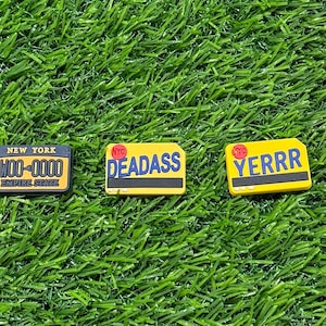 Könnte beinhalten: Drei gelbe und schwarze Schlüsselanhänger im Stil von Nummernschildern mit dem Text "NYC WOO-0000 EMPIRE STATE", "NYC DEADASS" und "NYC YERRR" darauf.