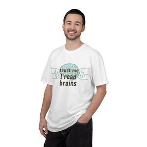 Könnte beinhalten: Weißes T-Shirt mit einer Abbildung eines türkisfarbenen Gehirns und dem Text "trust me I read brains" in Braun. Das T-Shirt hat kurze Ärmel und einen Rundhalsausschnitt.