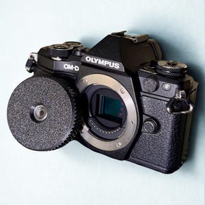 May include: A black Olympus OM-D camera with the lens cap removed, revealing the camera's sensor. The camera has a silver ring around the lens and the word "OLYMPUS" printed on the top.