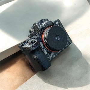 Peut inclure: Un appareil photo Sony Alpha 7R noir et gris avec un motif de camouflage et des accents orange. L'appareil photo a un capuchon d'objectif sur l'objectif.