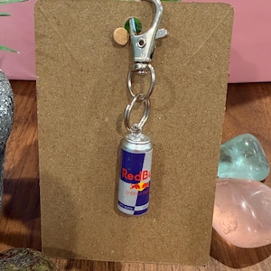 Op de afbeelding: Een zilverkleurige metalen sleutelhanger met een miniatuurreplica van een Red Bull energiedrankblikje. De bedel is bevestigd aan een zilveren karabijnhaak en een ring, gemonteerd op een rechthoekige bruine kartonnen displaykaart.