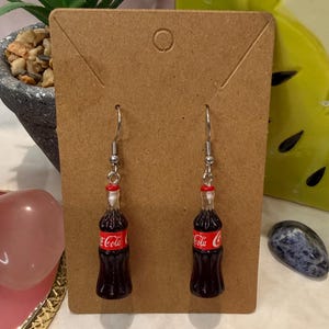 May include: A pair of dangle earrings featuring miniature dark brown glass-like soda bottles with red labels that read Coca-Cola. The earrings have silver-toned metal hooks and are displayed on a rectangular brown cardboard jewellery display card.