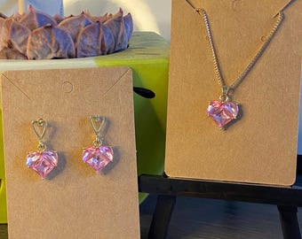 May include: A gold-toned necklace and earrings set. The necklace features a pink heart-shaped pendant. The earrings also have pink heart-shaped pendants. The jewelry is displayed on brown cardboard cards.