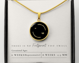 May include: A gold necklace with a black circular pendant featuring two tiny footprints. The pendant has the text "KATERINA LYNN" and "DAISY CARTER JOHNSON" inscribed around the edge. The necklace is in a black box with the text "THERE IS NO Footprint TOO SMALL" and "Gestational Ages: 6 WEEKS (Representative) 8 WEEKS 2.3 MM".