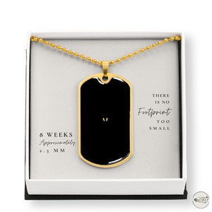 May include: Gold necklace with a black dog tag pendant. The dog tag has a small footprint image and the text "There is no footprint too small". The necklace is in a white box with the text "8 weeks approximately 2.3 mm" on the bottom.