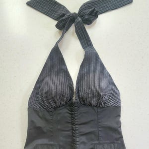 May include: A black halter top featuring a pinstriped bodice with a tie-neck closure and a solid black lower section. The garment includes a deep V-neckline, structured cups, and two faux welt pockets at the waist, creating a tailored appearance.