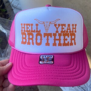 Könnte beinhalten: Pinke und weiße Trucker-Mütze mit dem Text "HELL YEAH BROTHER" in Orange. Die Mütze hat einen Mesh-Rücken und einen gebogenen Schirm. Das Design beinhaltet eine Longhorn-Schädel-Grafik.