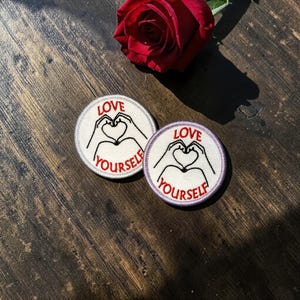 May include: Two round embroidered patches with the words "LOVE YOURSELF" and a heart formed by hands. The patches are white with black and red accents. A red rose is in the background.