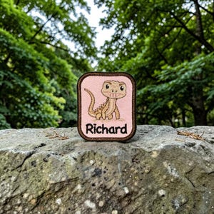 May include: A rectangular embroidered patch with a light pink background and a dark brown border. The patch features a cartoon lizard illustration and the name Richard printed in black text below the lizard. The patch has rounded corners.