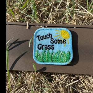 May include: A square, embroidered patch with a light blue background and a white border. The patch features the words "Touch Some Grass" in black, a yellow sun, and green grass. The patch is resting on a brown surface with grass in the background.
