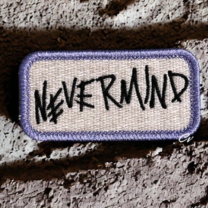 May include: A beige fabric patch with a purple border. The patch has the word "Nevermind" written in black script.