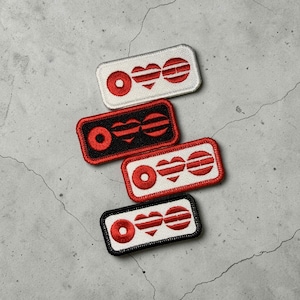 May include: Four rectangular embroidered patches with a red and white design. Each patch features a red circle, a heart, and two horizontal lines. The patches have black or white borders and are arranged on a gray surface.