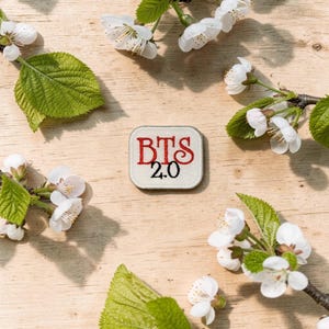 May include: A square patch with the text "BTS 2.0" embroidered in red on a cream background. The patch is surrounded by white cherry blossoms and green leaves, all arranged on a light wooden surface.