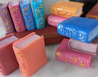 May include: A collection of decorative, miniature books with gold accents and intricate designs. The books are arranged in a stack and are made of a variety of colors, including pink, blue, and orange.