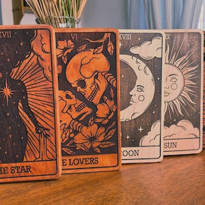 May include: A collection of wooden tarot cards featuring intricate designs and illustrations. The cards depict imagery such as a star, lovers, a moon, a sun, and a devil. The cards are rectangular with dark brown engraved artwork on a lighter brown background.