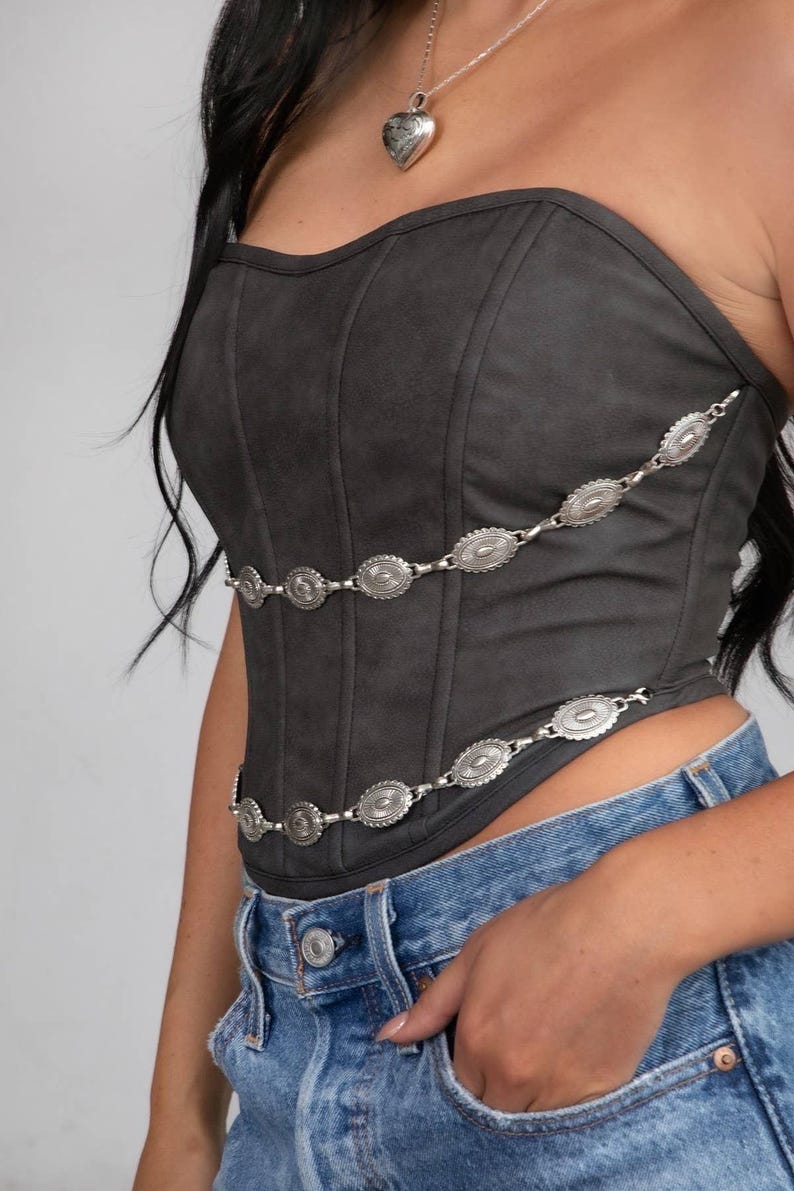May include: A dark gray corset top with silver-colored chain detailing. The corset has a structured, fitted design and is paired with blue denim jeans. A silver heart-shaped pendant hangs from a necklace.