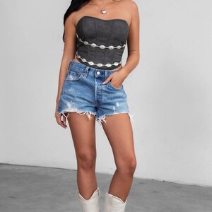 May include: A dark gray strapless corset top with silver embellishments is paired with distressed blue denim shorts. The outfit is completed with white cowboy boots, creating a stylish, casual ensemble.