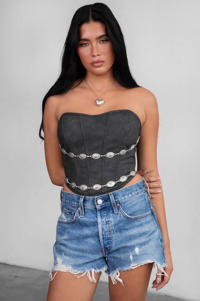 May include: A dark gray strapless corset top with silver embellishments and a matching silver heart pendant necklace. The top is paired with blue denim shorts.