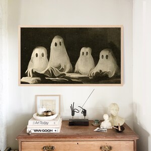 Puede incluir: Una impresi&oacute;n enmarcada representa cuatro figuras de fantasmas blancos con ojos negros sobre un fondo oscuro. La obra de arte cuelga sobre un mueble de madera con objetos decorativos, incluyendo libros, una vela y peque&ntilde;as esculturas. El mueble tiene dos cajones y dos puertas.