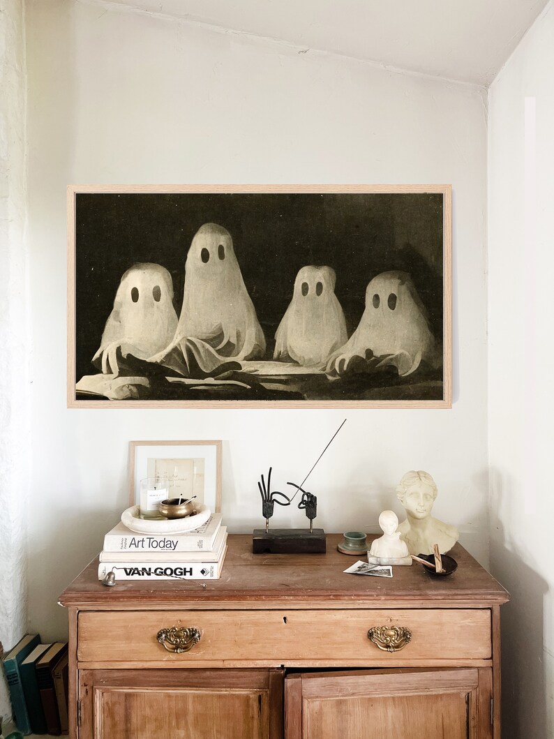 Puede incluir: Una impresi&oacute;n enmarcada representa cuatro figuras de fantasmas blancos con ojos negros sobre un fondo oscuro. La obra de arte cuelga sobre un mueble de madera con objetos decorativos, incluyendo libros, una vela y peque&ntilde;as esculturas. El mueble tiene dos cajones y dos puertas.