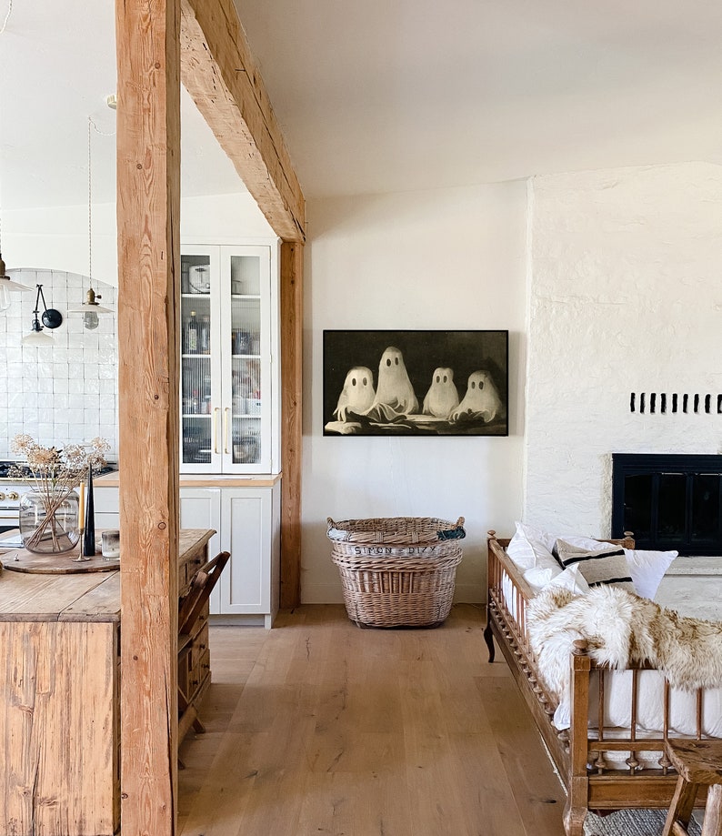 Puede incluir: Una obra de arte enmarcada con cuatro figuras de fantasmas blancos sobre un fondo oscuro cuelga de una pared blanca. Una gran cesta de mimbre descansa sobre el suelo de madera. La habitaci&oacute;n tiene una est&eacute;tica r&uacute;stica de granja con vigas de madera vistas y una chimenea.